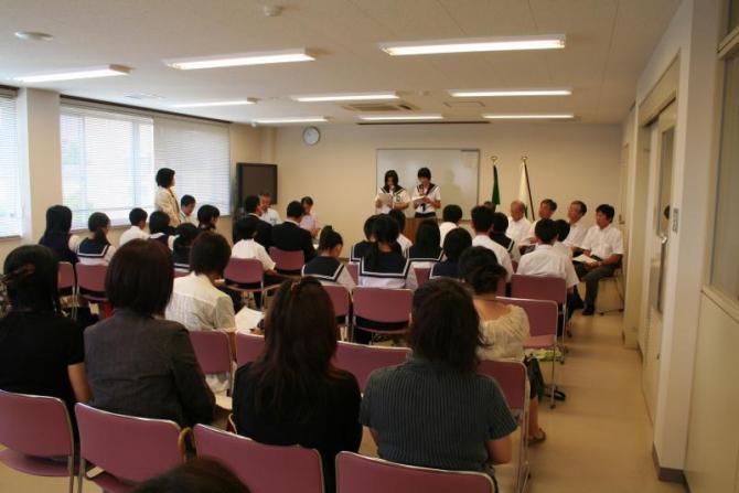 教室の前方に立ち、フランス語によるあいさつをしている2人の女子生徒とあいさつを聞いている生徒たちと職員の写真