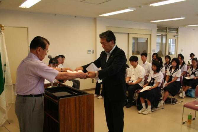 教室前方の演壇に立ち団長に親書を手渡している町長とその様子を椅子に腰かけて見ている生徒たちの写真