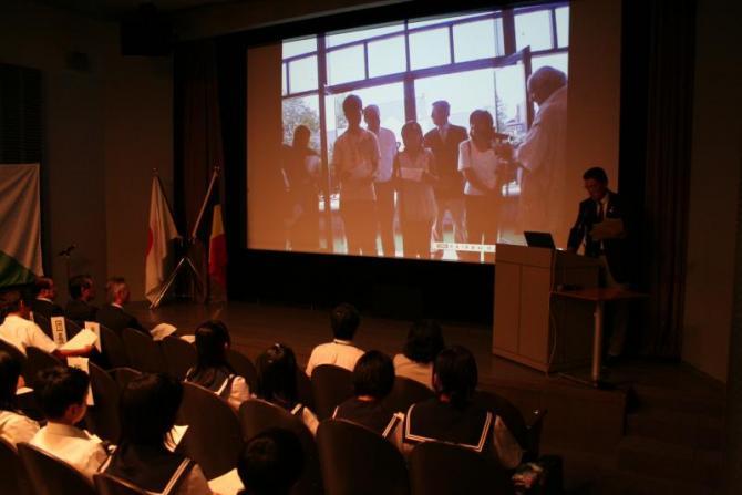 スクリーンの横に立ち訪問の報告をしている引率者のと話を聞いている生徒たちの写真