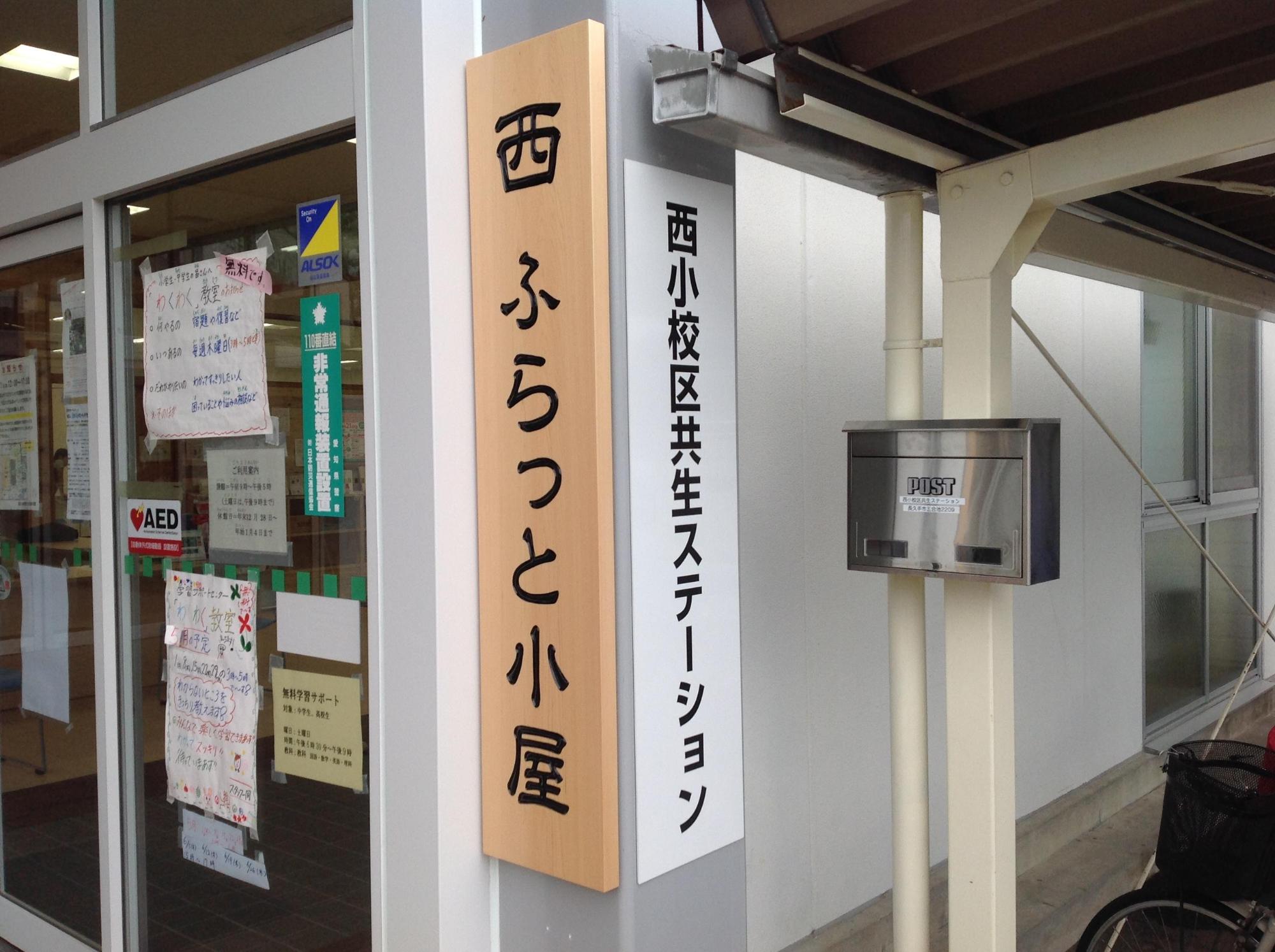 建物の入り口の右側に、西小校区共生ステーション「西ふらっと小屋」と書かれた看板が設置されている写真