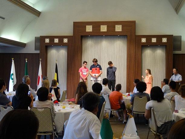 前方の舞台に立って挨拶をしているワーテルロー市訪問団の3人の学生と席で見守っているお別れ会の参加者の写真
