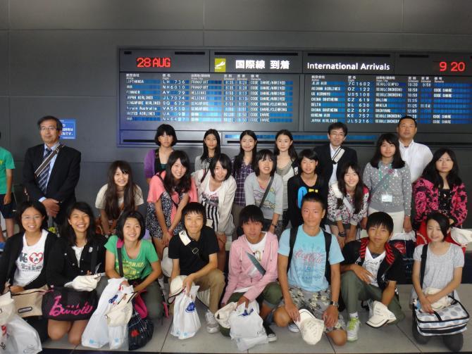 空港の国際線のフライト案内表の前で参加者が集まって笑顔で写っている集合写真