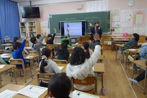 講師の先生がテレビに映像を流して話をしており、席についている児童が右手を挙げて挙手をしている授業風景の写真