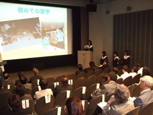 スクリーンに映しだされた映像を見ながら発表をしている女子生徒と発表を聞いている来場者の写真