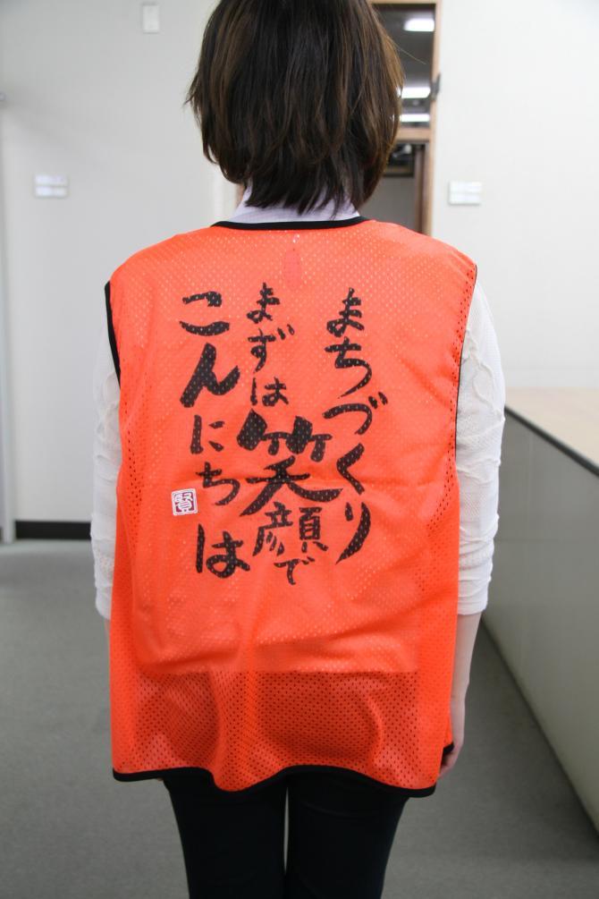「まちづくり まずは笑顔でこんにちは」と書かれたオレンジ色のあいさつ運動啓発ベスト背面