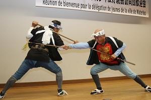 ハチマキを頭につけて、タスキがけをした衣装を身に着けた男性2名が棒を持って向かい合い、左脚を引いて棒を交差させ棒の手を披露している写真