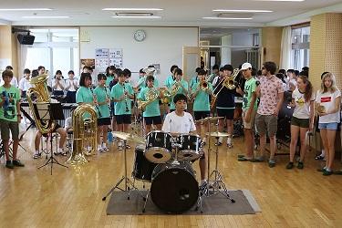 中央にドラムの演奏、ドラムの後ろにクラリネットやホルン、トランペット、トロンボーンなどの楽器を演奏をしており、右側に立っている訪問団の方々が演奏を聴いている写真