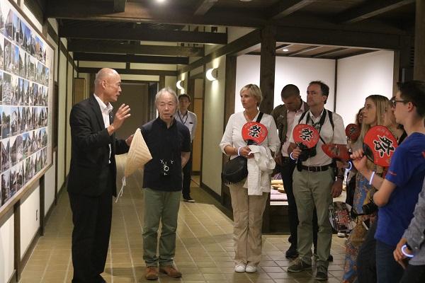 壁にたくさんの写真が展示してあり、祭りと書かれた赤いうちわを持っている訪問団の方々に黒いスーツを着た男性が説明をしている写真