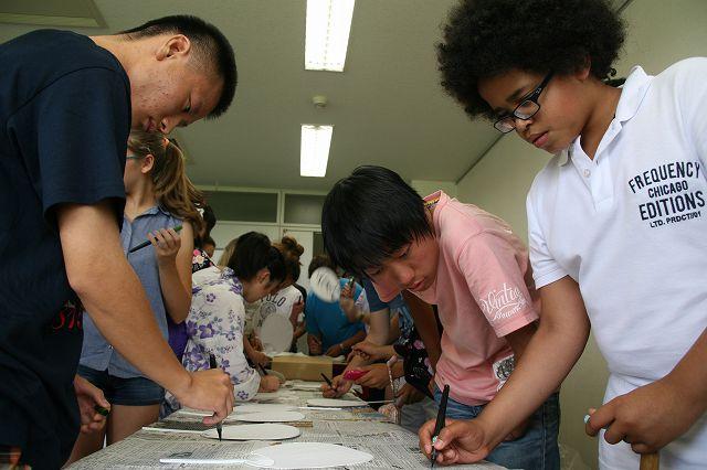うちわにペンで模様を描いている、長久手市の男子学生とワーテルロー市の男子学生の写真