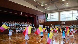 体育館のステージの上に整列したマーチングバンドが演奏をした曲に合わせて、ステージ下のカラーガード隊が三色の旗を振って演技している写真