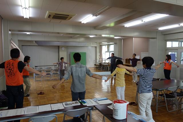 前に立っている女性が両手を広げているのを、間隔を開けて立っている参加者が見ながら同じポーズをとっている写真