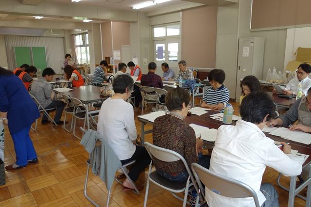 2つの長机をくっつけて6か所並べ、それぞれパイプ椅子に参加者が座って、配られた資料を熱心に見ている写真