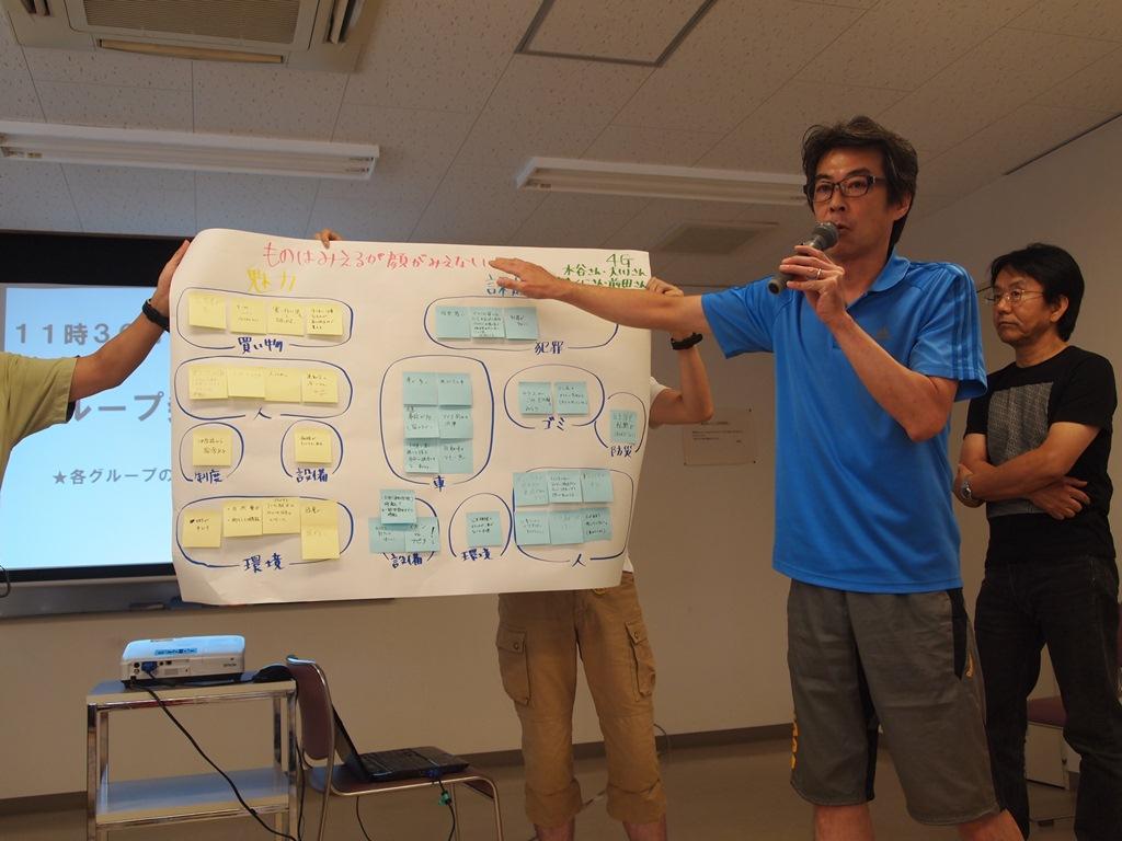 青いTシャツを着た男性が作成した資料を手で指し、マイクを持ちながら説明している写真