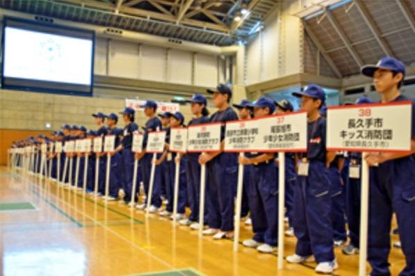 少年消防クラブ交流会にてプラカードを持ったキッズ団員を先頭に整列している写真