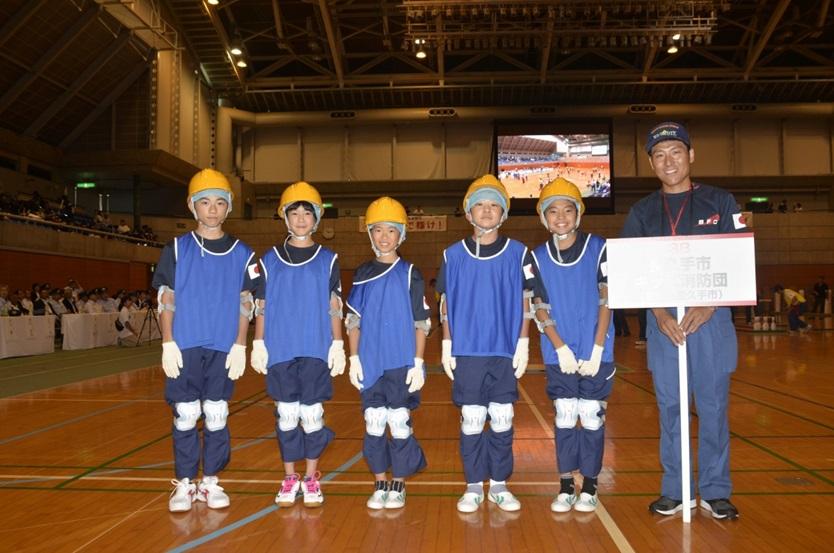 プラカードを持った消防団員の横に黄色いヘルメットに青いベストを着用したキッズ消防団員5名が並んで記念撮影している写真
