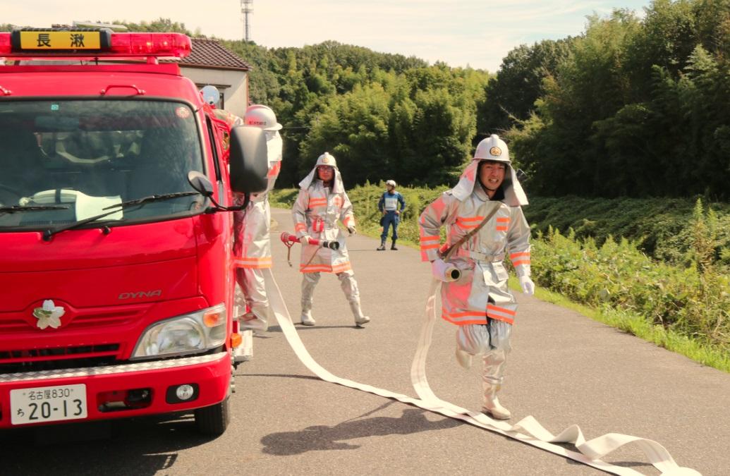 白い消防服を着た団員たちが昼間に民家の近くでホースを持って火災訓練をしている写真