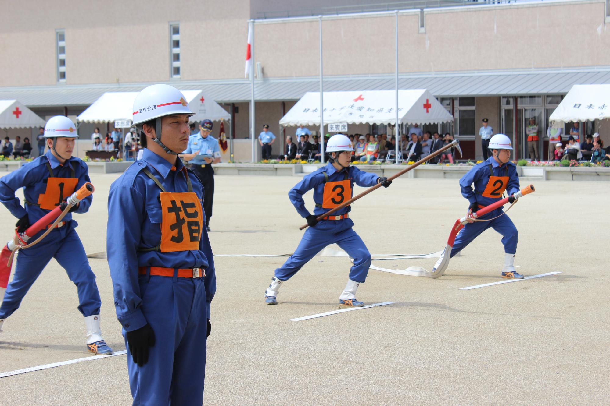 消防操法大会にて指揮者が一番先頭に立ち、後ろにゼッケン1と2の消防団員が放水ホースを持ち、1と2の間にゼッケン3の消防団員がかぎの付いた棒を持って等間隔に立っている写真