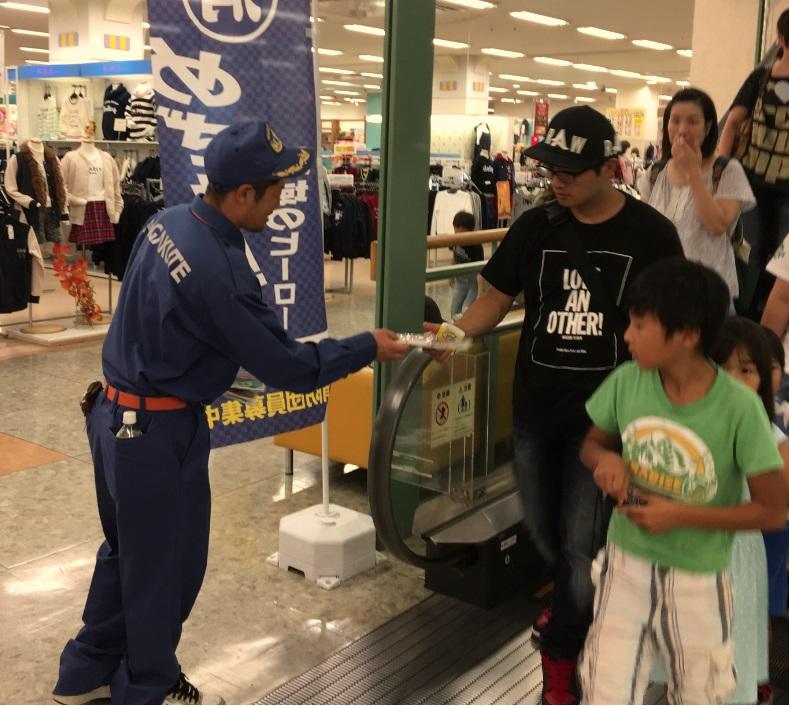 入団募集ののぼり旗を設置したスーパーのエスカレーターの降り口で来店者に消防団入団募集の資料を配る消防団員の写真