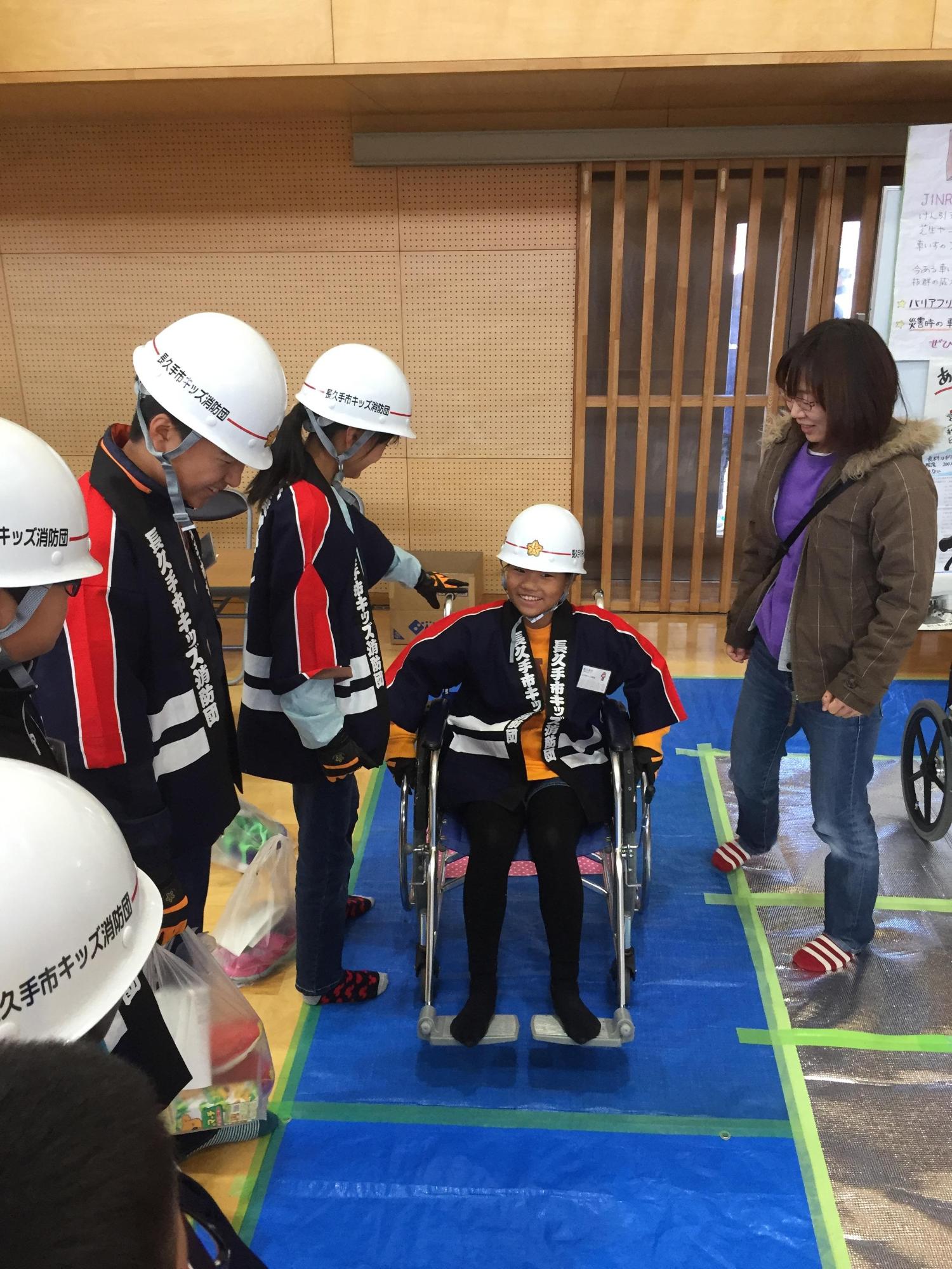 ヘルメットに法被姿のキッズ消防団員が体育館で車いす体験をしている様子を他のキッズ消防団員が見ている写真