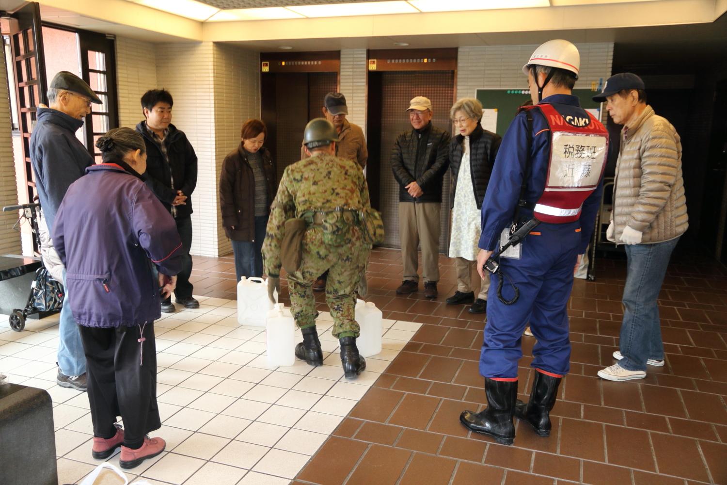 マンションのロビーのエレベーター前で給水タンクを陸上自衛隊員が置いている周りで住民やヘルメットを被った市職員が説明を聞いている写真