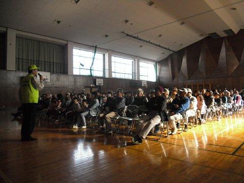 体育館内の前方だけカーテンを引いて薄暗くした中に、設置したパイプ椅子に参加者が座っている前で、男性スタッフがマイクを片手に話しをしている写真