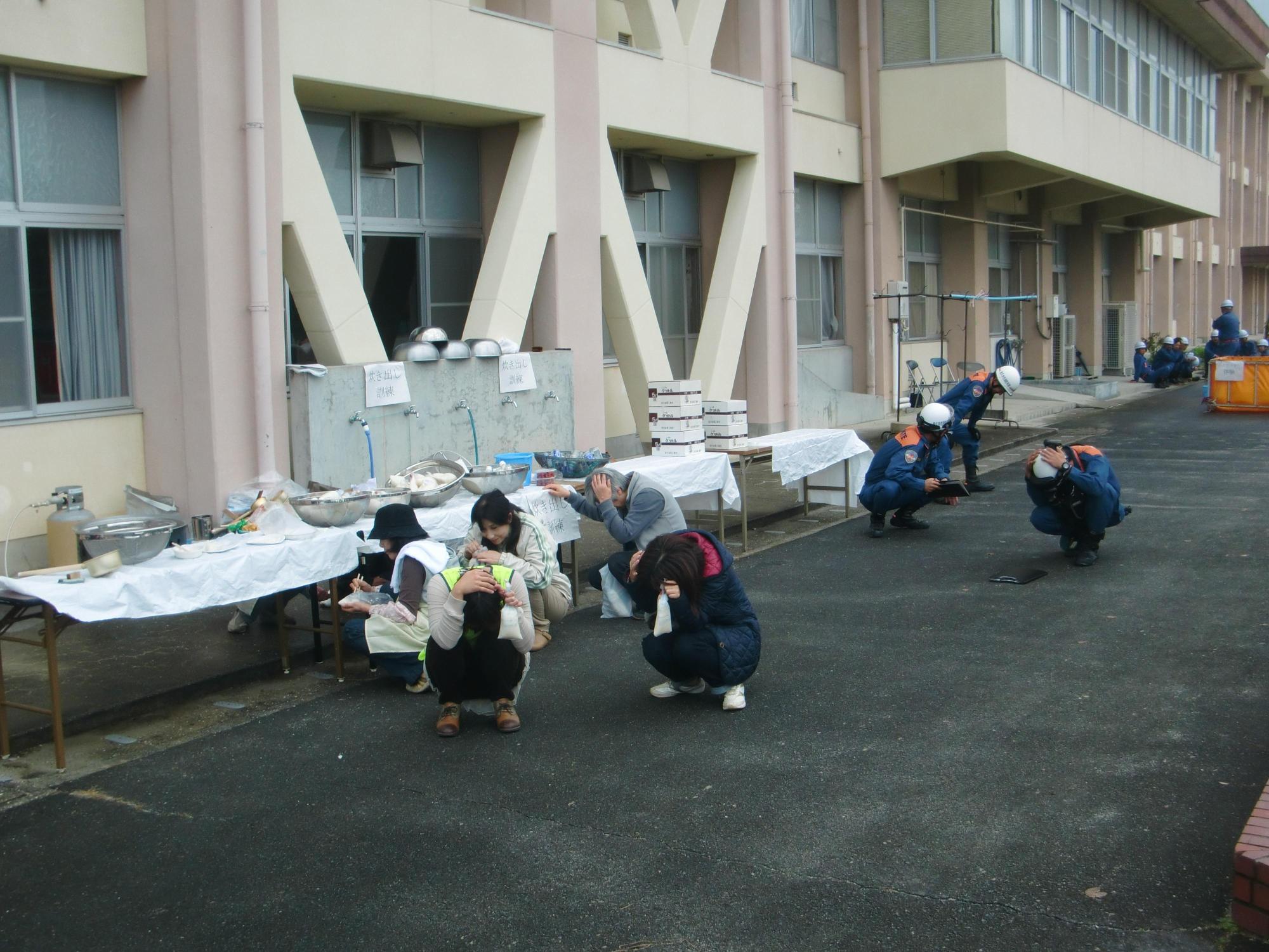 体育館の外の炊き出しブースの前で、消防団員や参加者たちが手を頭にのせうずくまっている写真