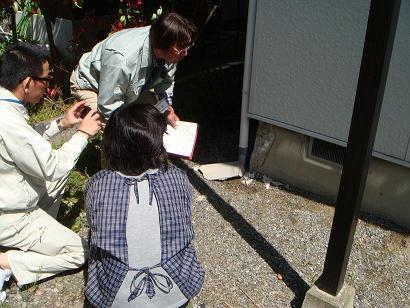 家の基礎部分にひび割れが入っており、エプソンを着た女性と市の職員の男性被害の箇所を見ており、もう一人の市の職員の男性が被害にあった場所をカメラで撮影している写真