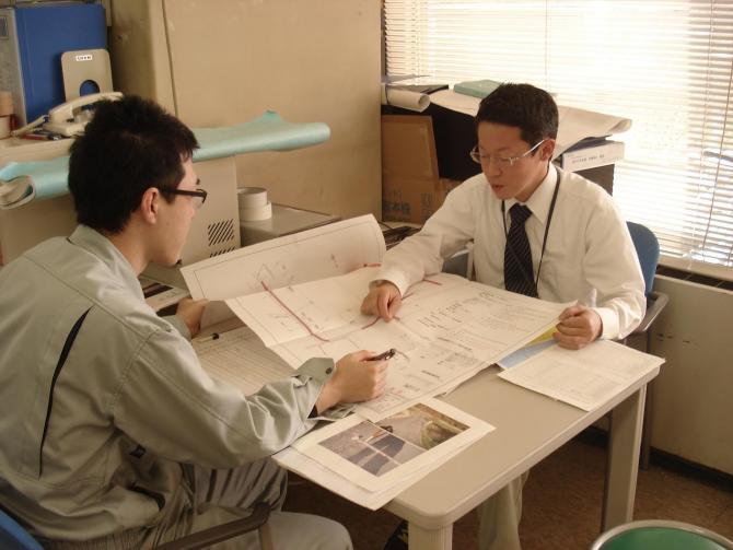 机に男性2名が向かい合って座っており、机の上に資料を広げて、話し合いをしている写真