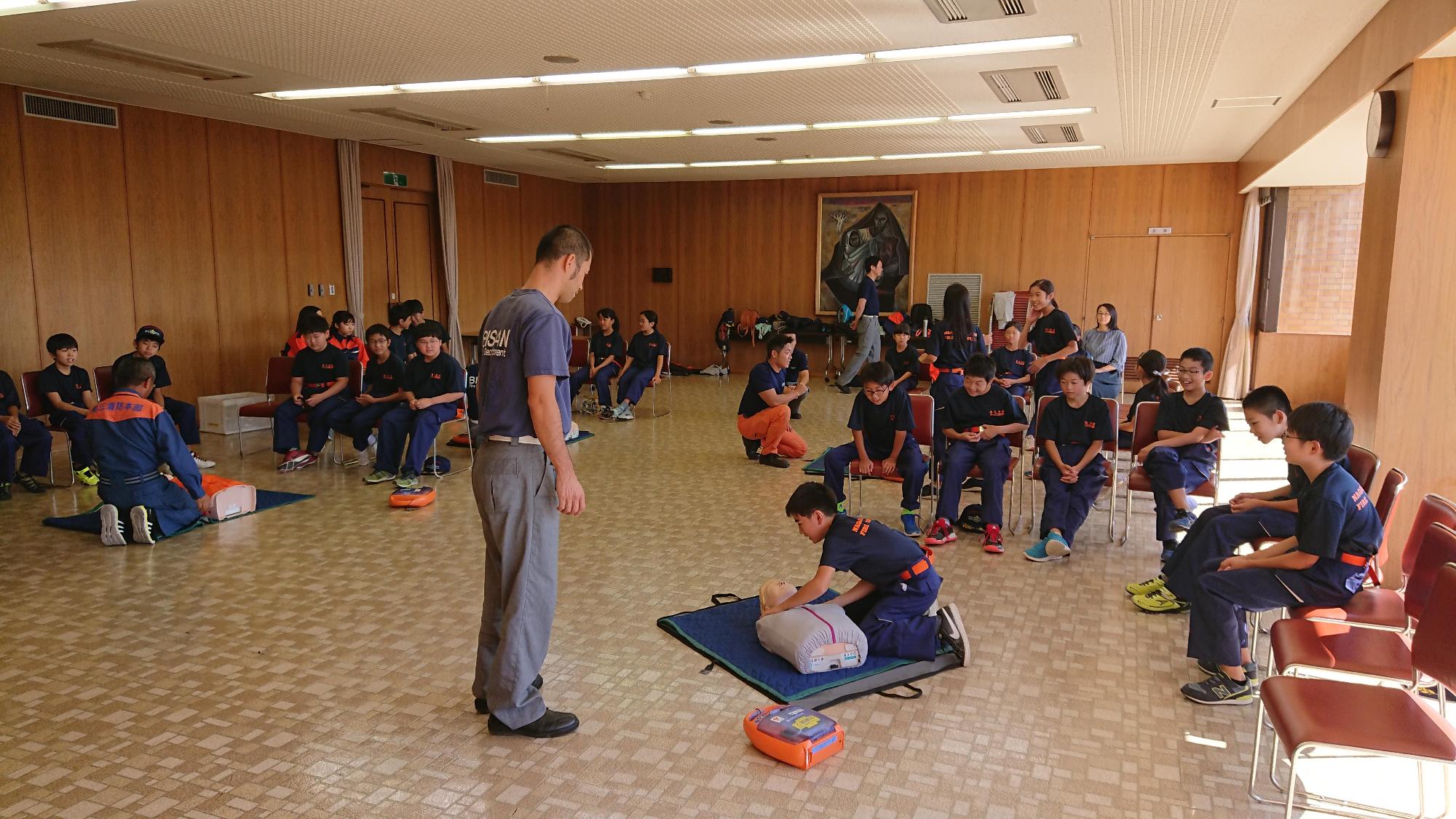 長久手市キッズ消防団員がグループごとに分かれて、講師からマネキンを使って心肺蘇生法を教わっている男子の様子を同じグループのメンバーが見ている写真