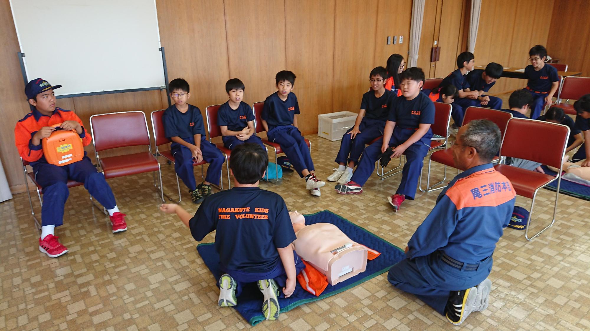 救命講習でマネキンを使って心肺蘇生法を教わっている男子が椅子に座っているメンバーにAEDをもらおうとしている様子を同じグループのメンバーと講師が見ている写真