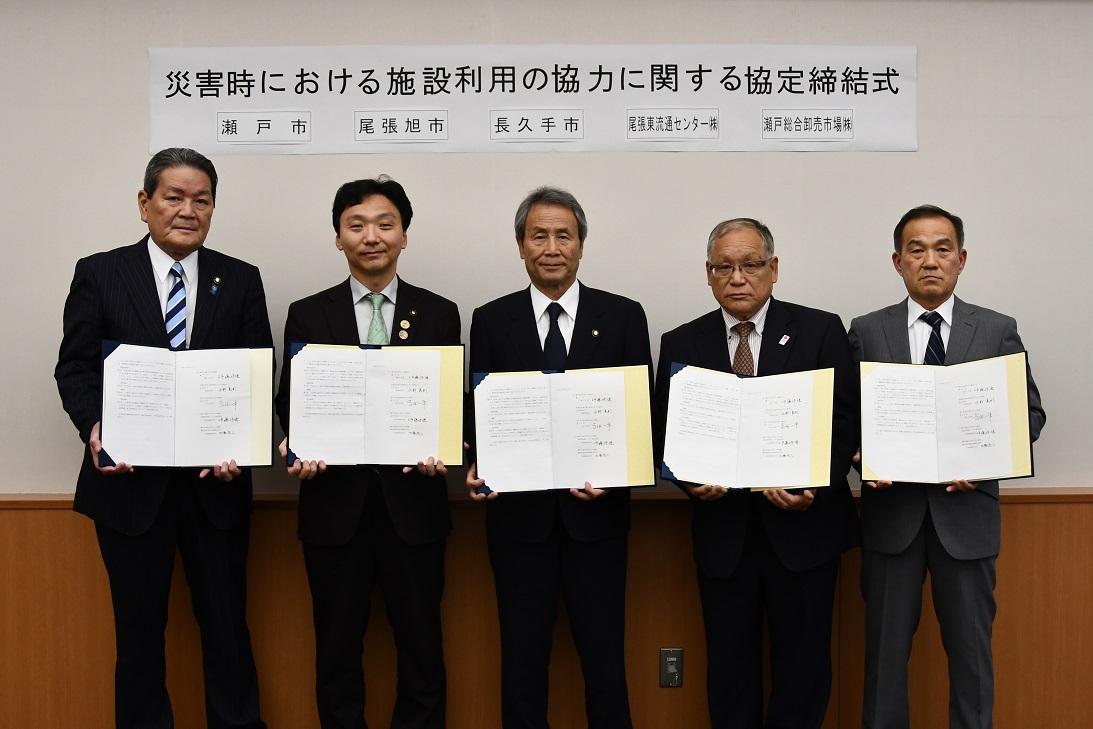スーツを着た5名の関係者たちがそれぞれ手に協定書を持ち記念撮影をしている写真