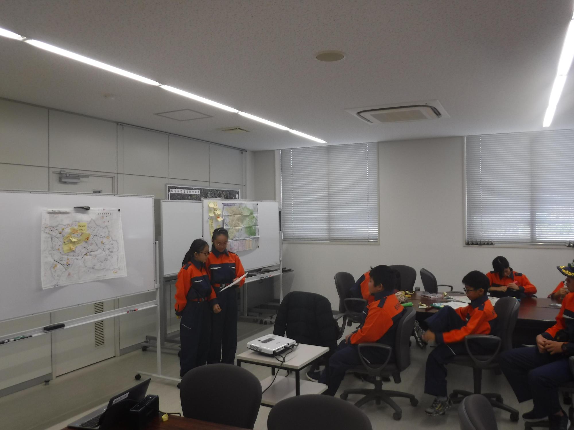 ホワイトボードに付箋やシールの貼られた地図が掲示されている前で、キッズ消防団員女子2名がみんなの前で調べたことを発表している写真