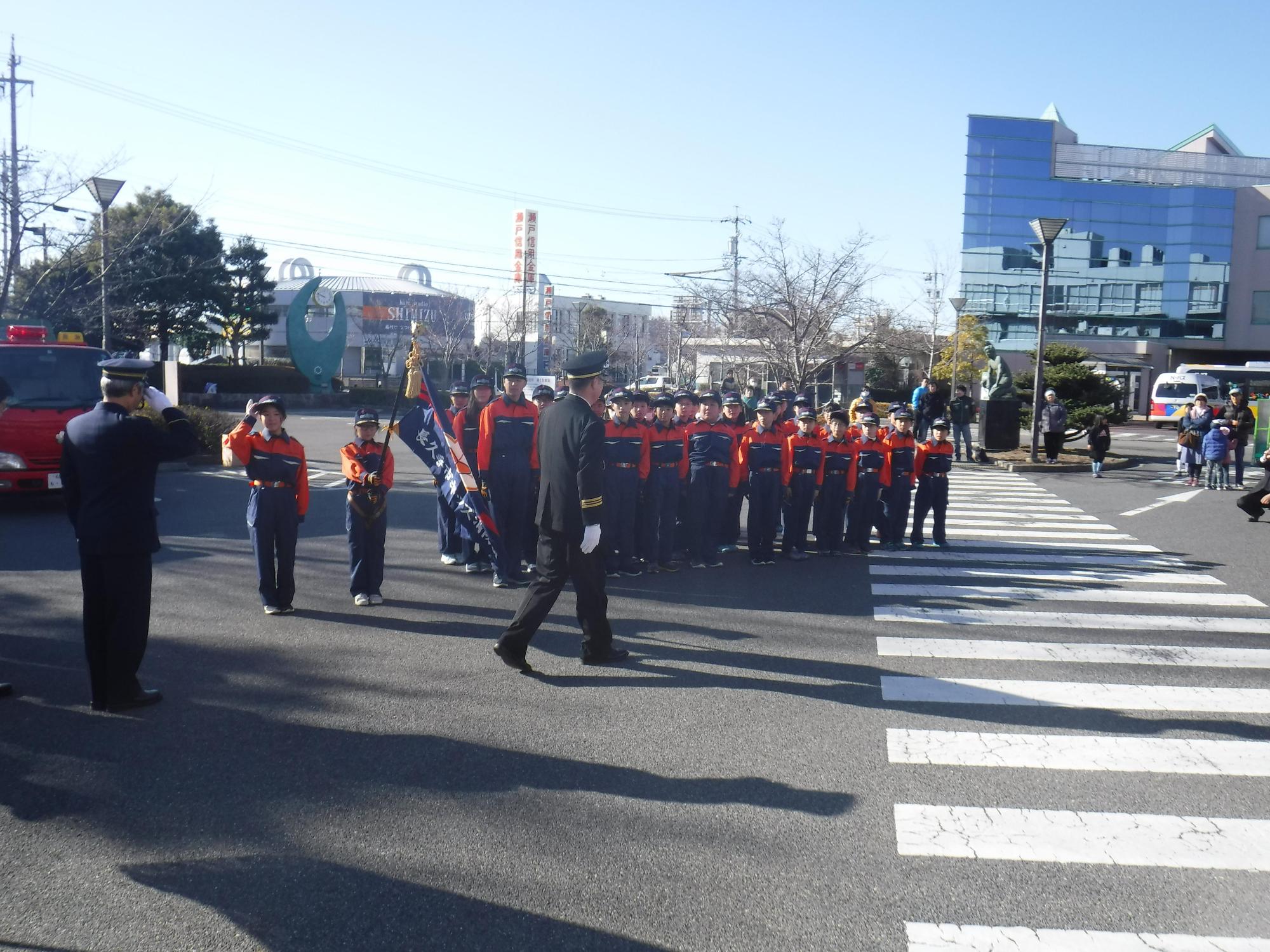 屋外で整列している参加者達と、そのうちの1人が制服を着た消防団員に敬礼をしている観閲式の様子の写真