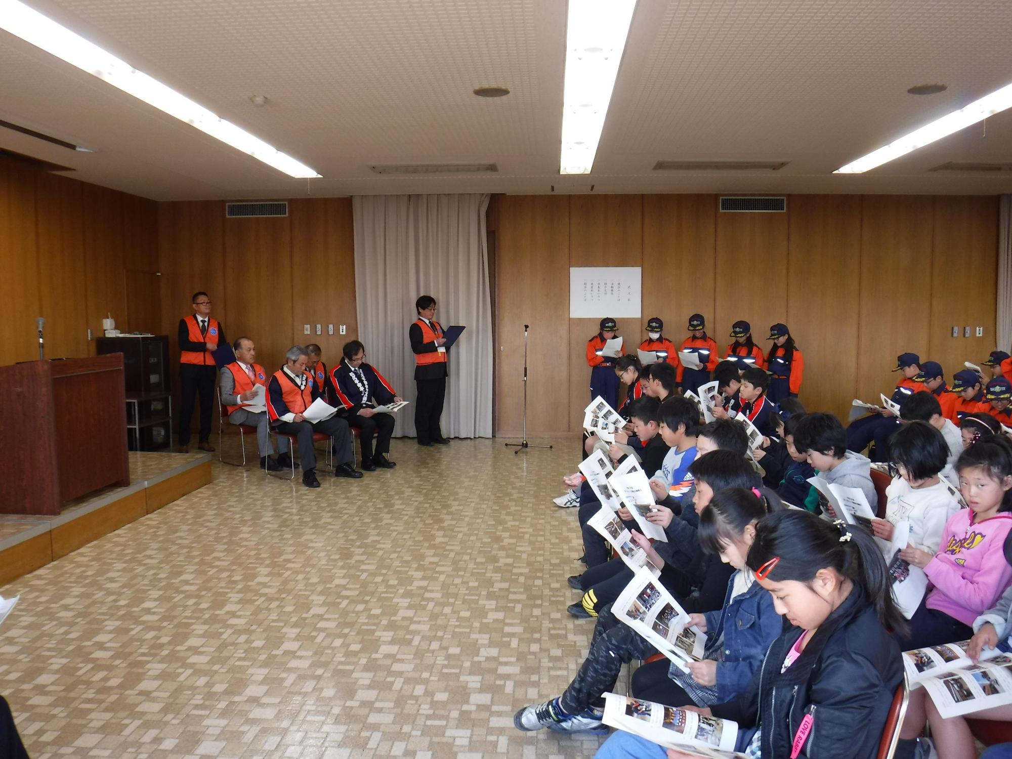 入退団式にて市長たちやスタッフ、私服姿の子供たち、消防団の制服姿のキッズ団員が椅子に座って、資料に目を通しながら司会者の話を聞いている写真