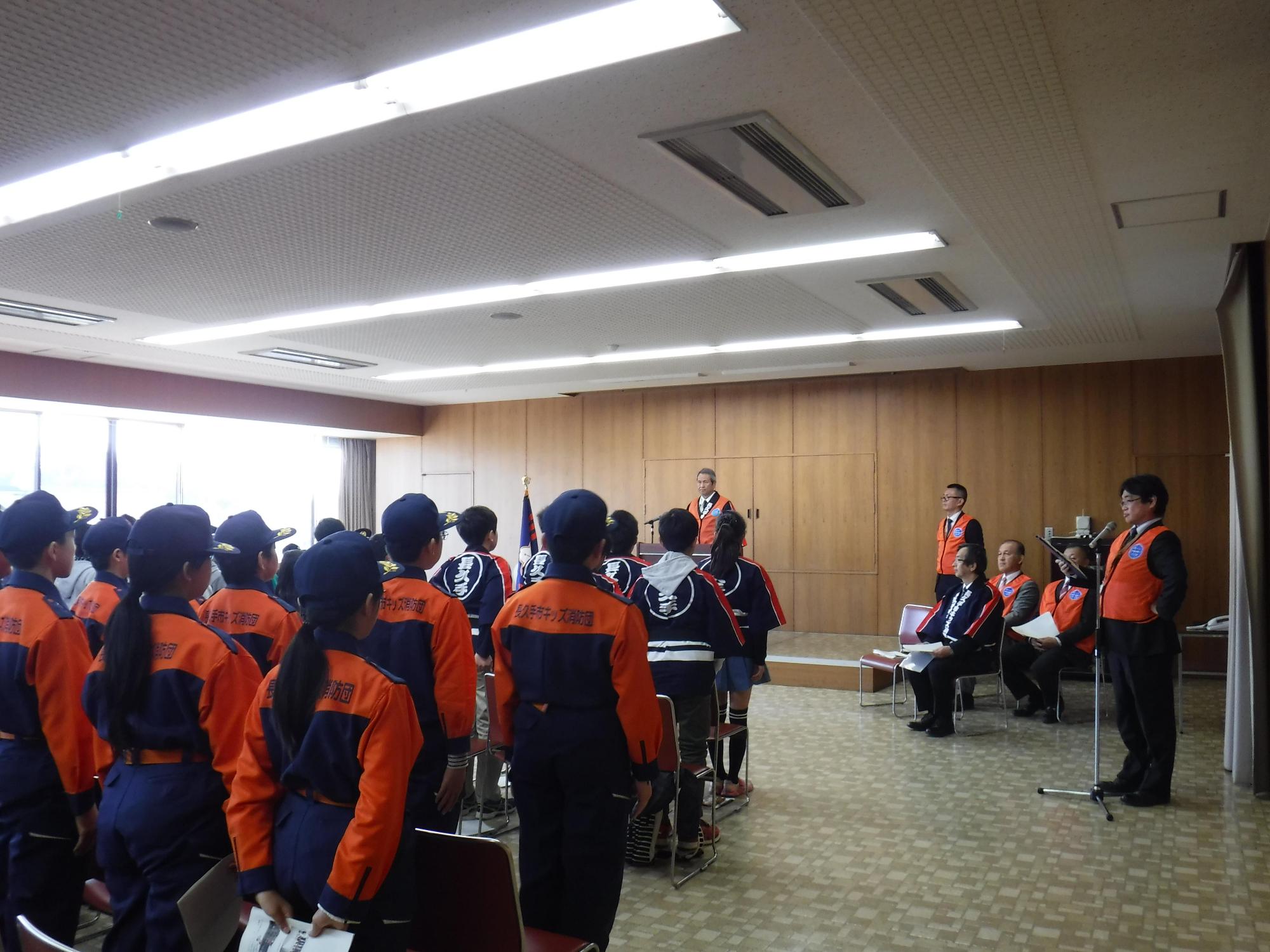 入退団式にて市長の前で入団した子供たち、消防団の制服姿や法被姿のキッズ団員たちが立って整列している写真