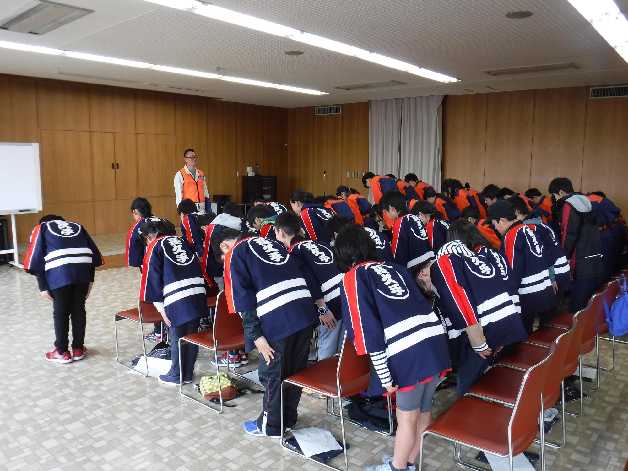 講堂の前の方でオレンジ色のベストを着た消防団員が立っており、後ろに並んでいる法被姿とユニホーム姿のキッズ消防団員が一同礼をしている写真