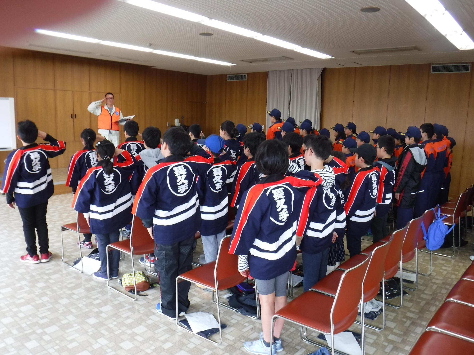 講堂の前の方でオレンジ色のベストを着た消防団員と、後ろに立って並んでいる法被姿やユニホーム姿のキッズ消防団員たちが敬礼している写真