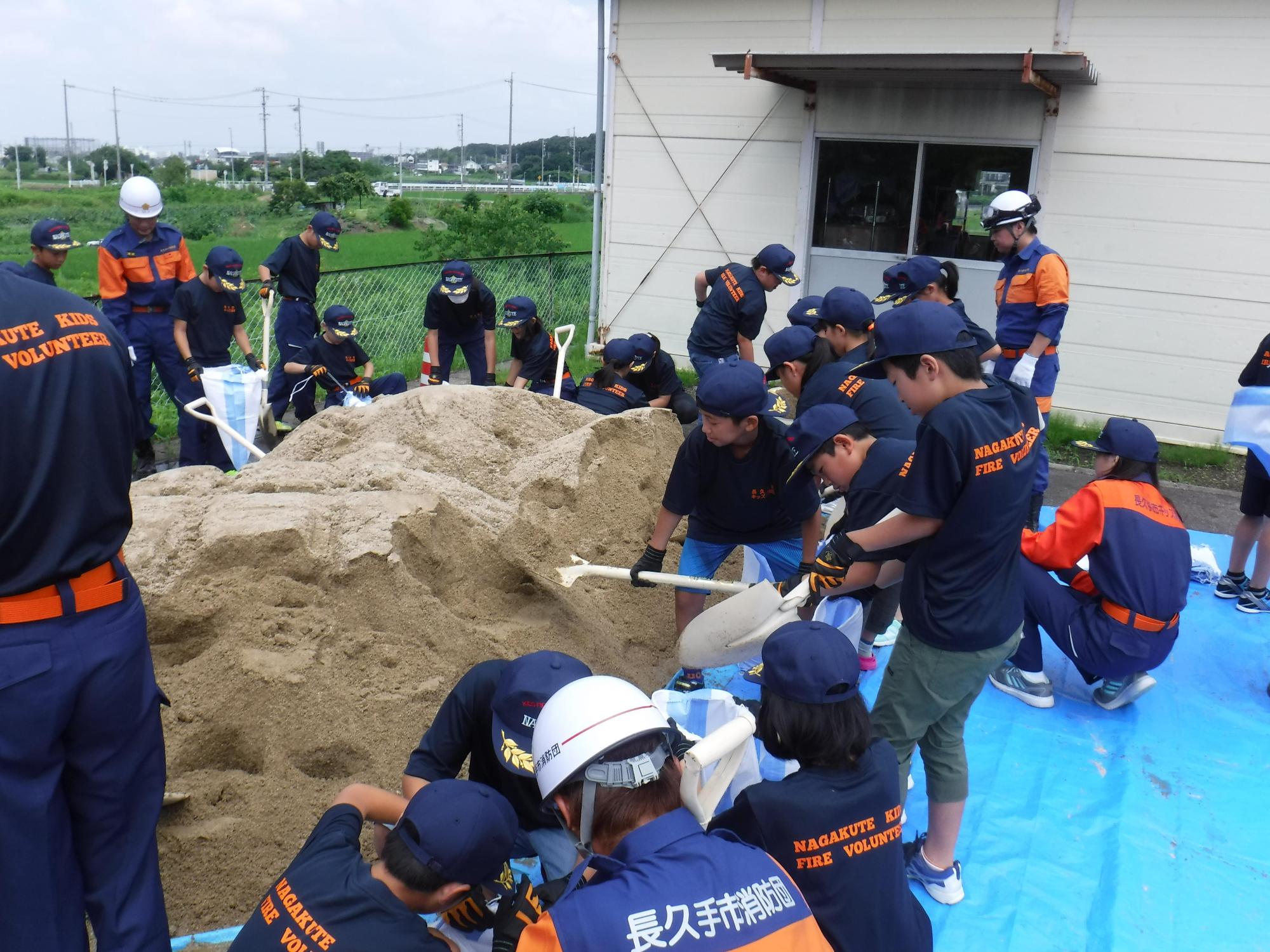 プレハブ倉庫入口前で消防団員の指導のもと長久手市キッズ消防団員たちがブルーシートの上の砂をスコップですくっている写真