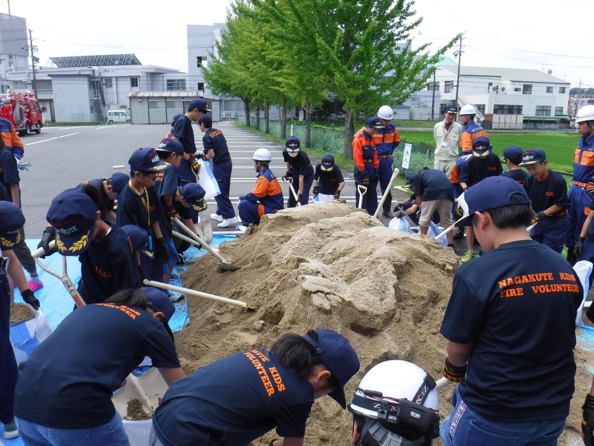 消防団員の指導のもと長久手市キッズ消防団員たちがブルーシートの上の砂をスコップですくっている先に駐車場と大きな木や建物が写っている写真