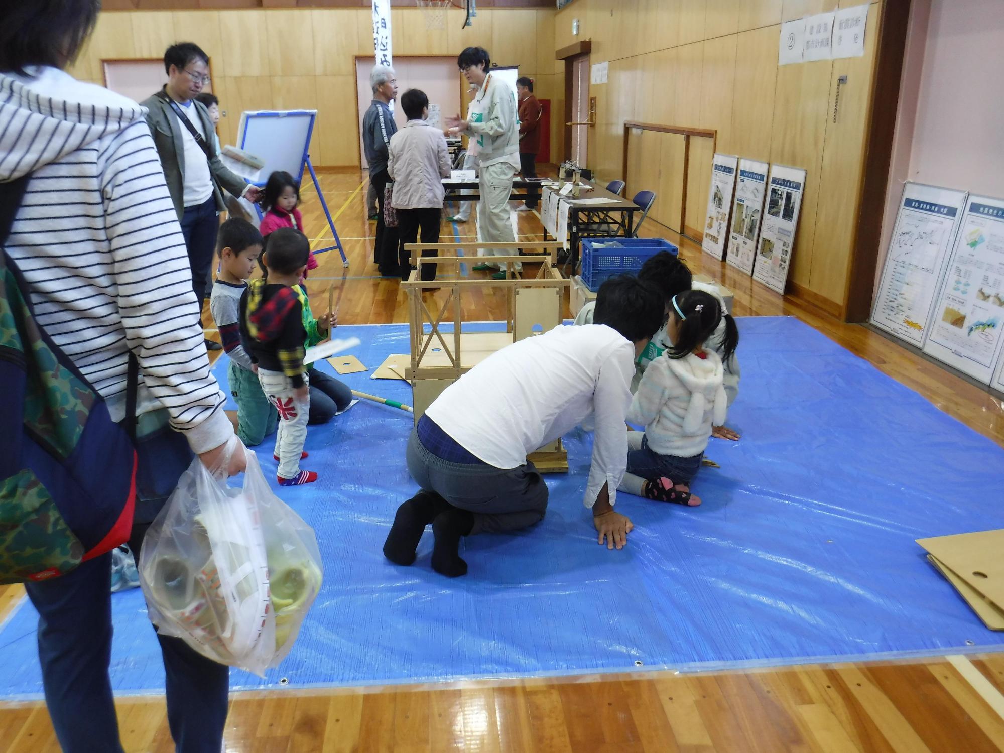 家の骨組みの模型や耐震診断のパネルが置かれているブースを親子たちが見学している様子と、奥で参加者夫婦が職員と話をしている写真