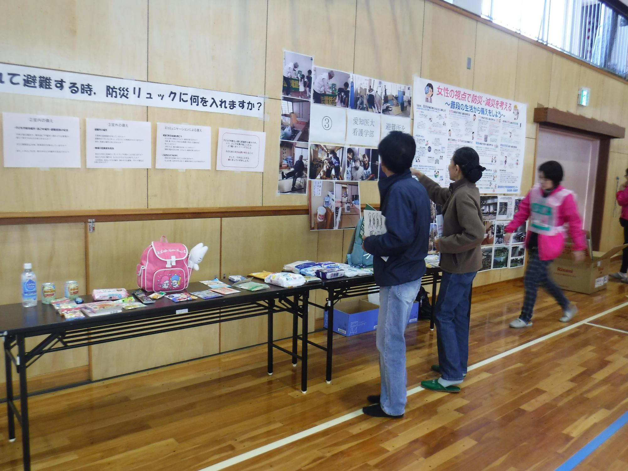 体育館の壁に避難所に関する資料が掲示されているブースに子供用のリュックや防災時にリュックに入れる中身が長机に展示されているのを見ている参加者たちの写真