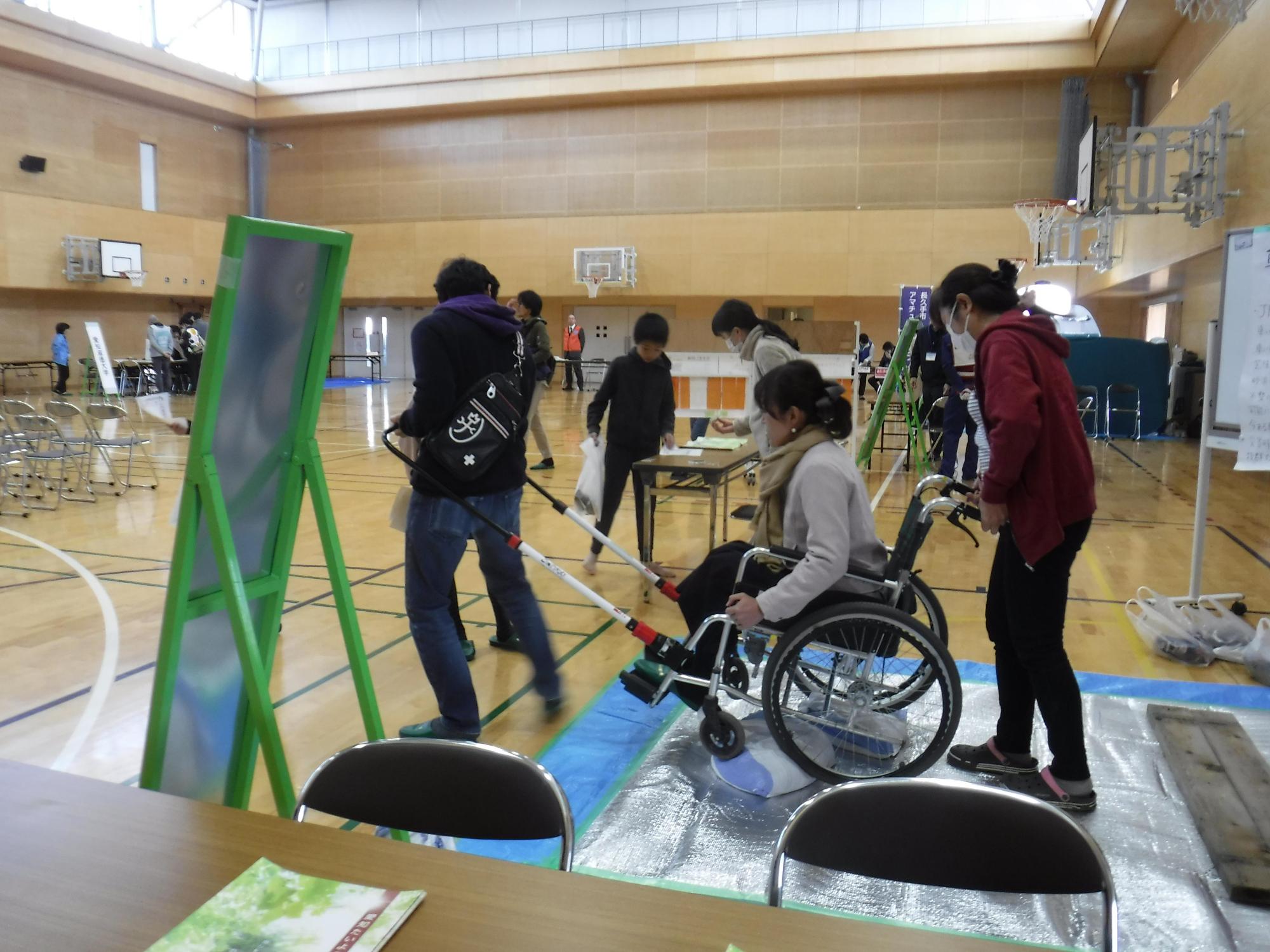 体育館に置かれた車いすで車いすを引く人、車いすに乗る人、車いすを押す人がそれぞれ体験している写真