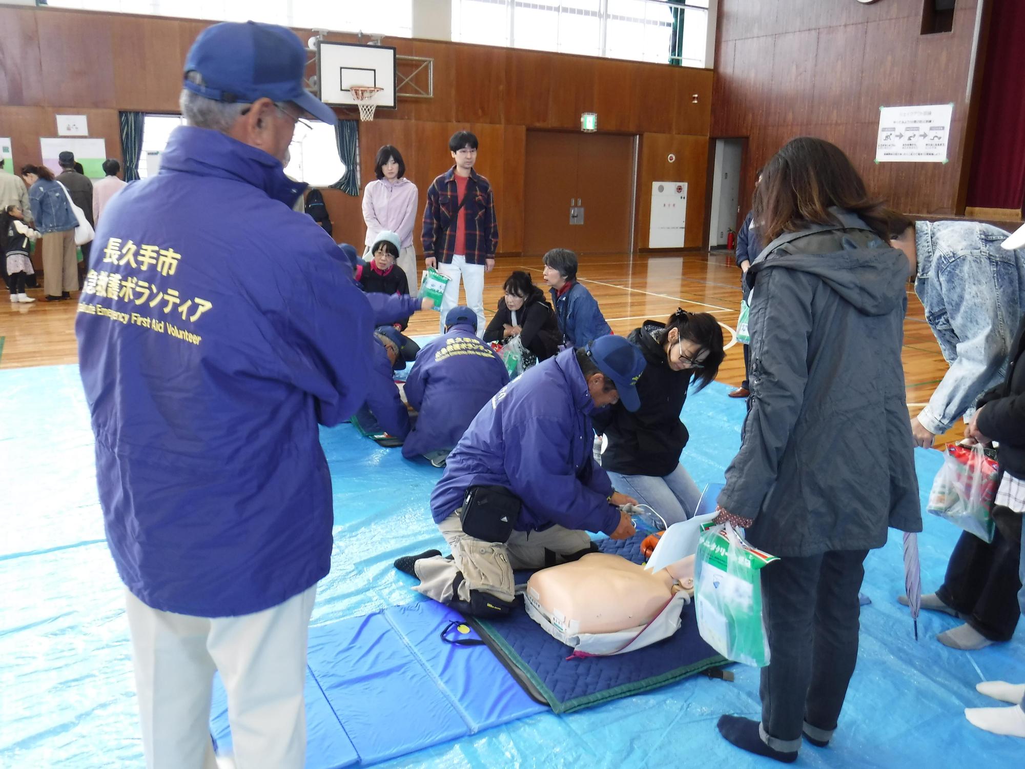 体育館に敷かれたブルーシートの上に救急救命用の人形があり、青色のジャンバーを着たボランティアスタッフに説明を受けながら訓練を受けている参加者たちの写真