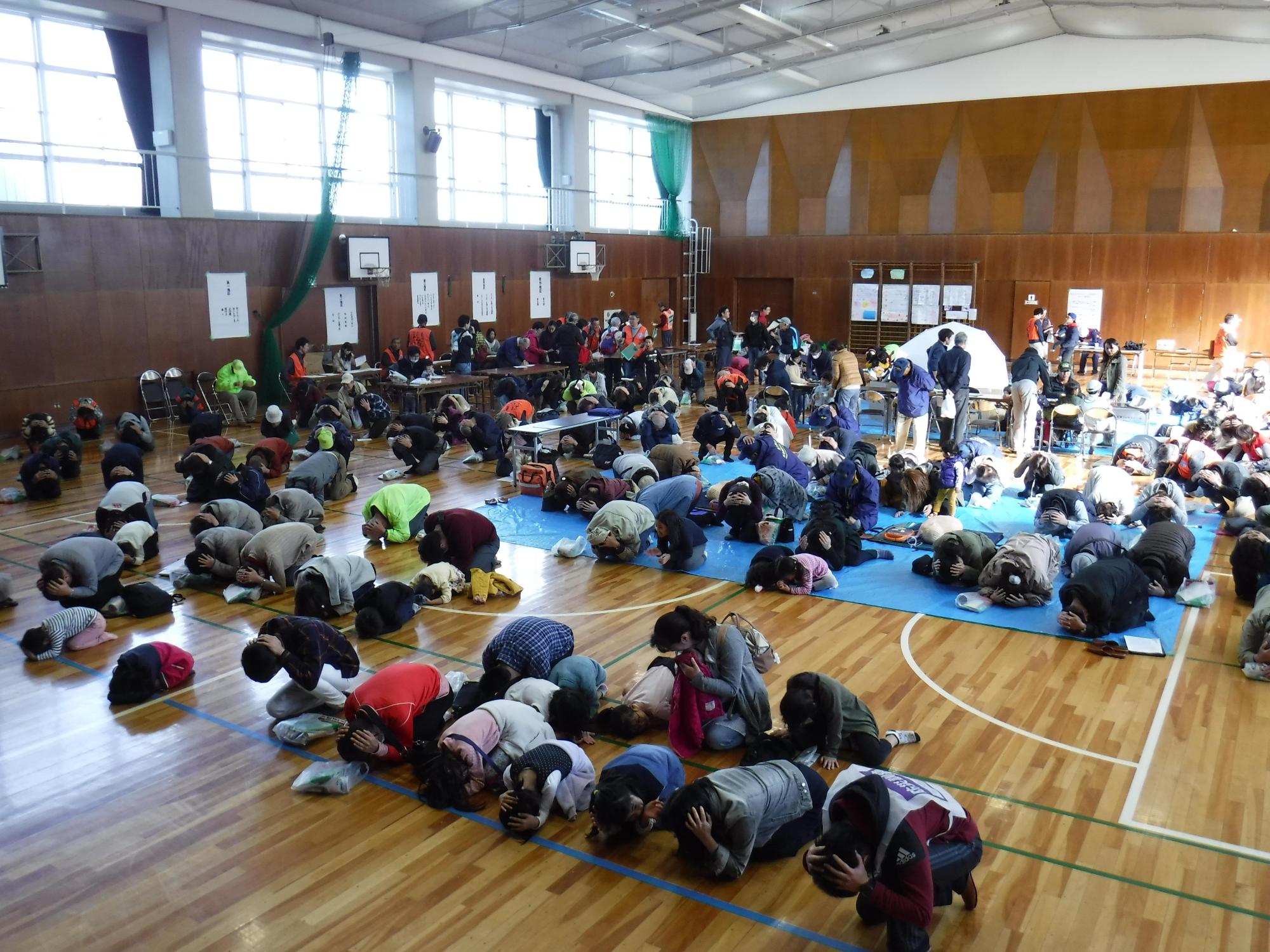 体育館で参加者たちが頭に手を添えて姿勢を低くしている(シェイクアウト訓練)写真
