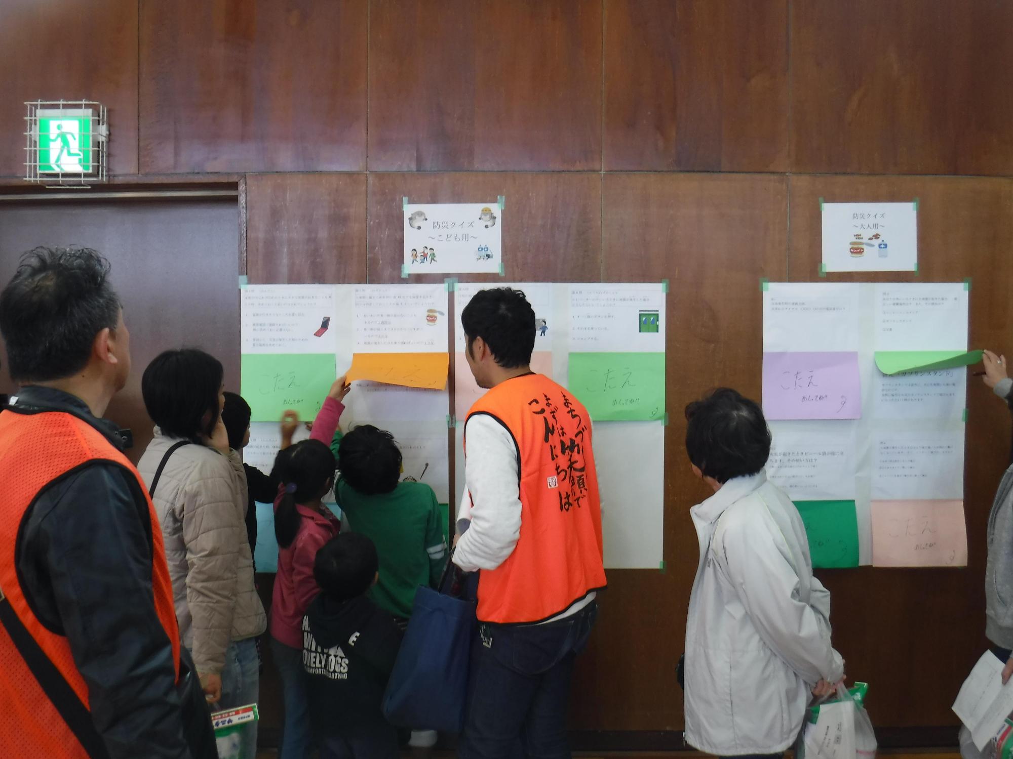 体育館に掲示された防災クイズに目を通す参加者や参加者の反応を見ているオレンジ色のベストを着たスタッフたちの写真