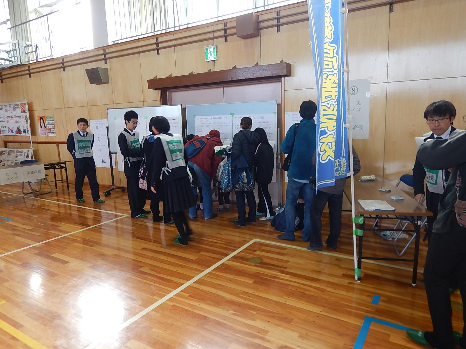 体育館内のホワイトボードに掲示された防災クイズに見入る参加者の周りに栄徳高等学校の学生たちが立っている写真