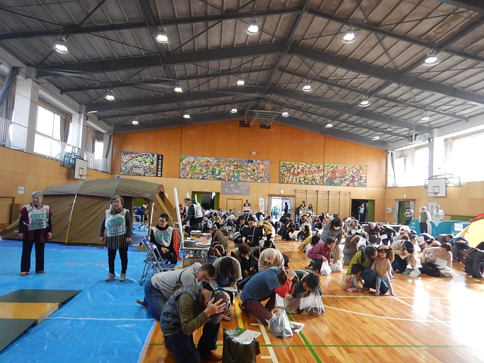 体育館で参加者たちが手を頭に添えて姿勢を低くしている(シェイクアウト訓練)写真