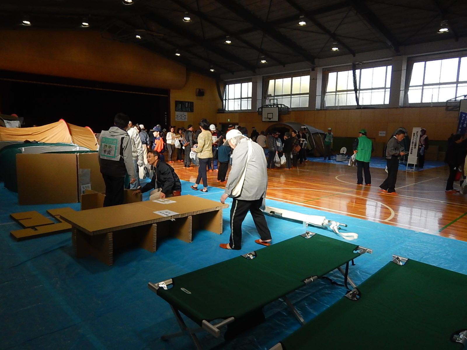 体育館に敷かれたブルーシートの上に段ボールベッドやテントなどの展示コーナーを見て回る人々の写真