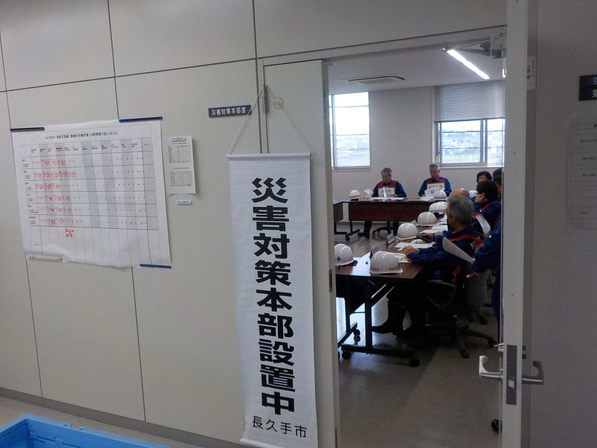 「災害対策本部設置中」ののぼり旗が本部の入口にかけられ、入口の戸が開いて対策本部員が打ち合わせしている様子が見えている写真