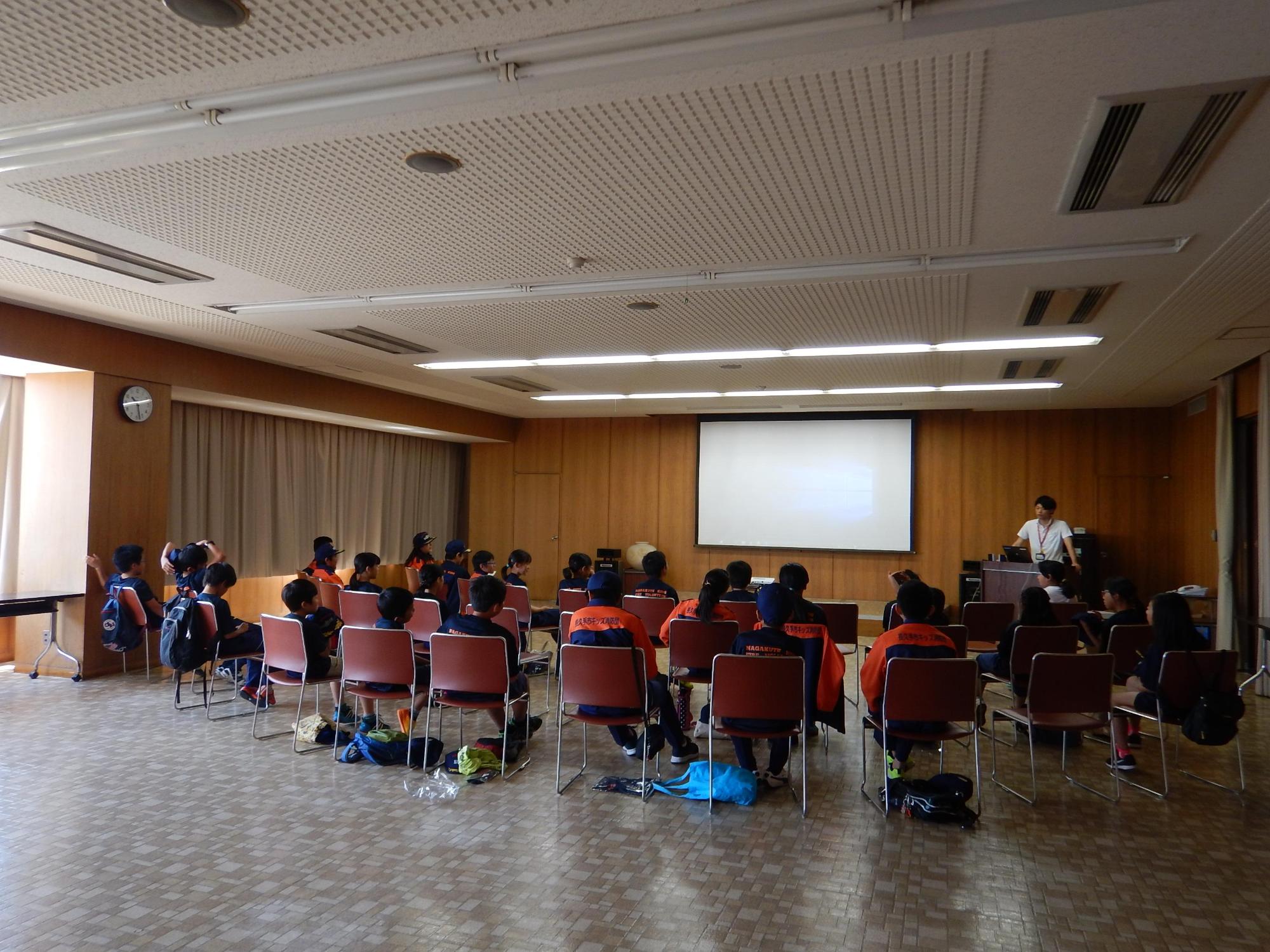 スクリーンの横で講師が話しているのを聞いているキッズ消防団員たちの写真